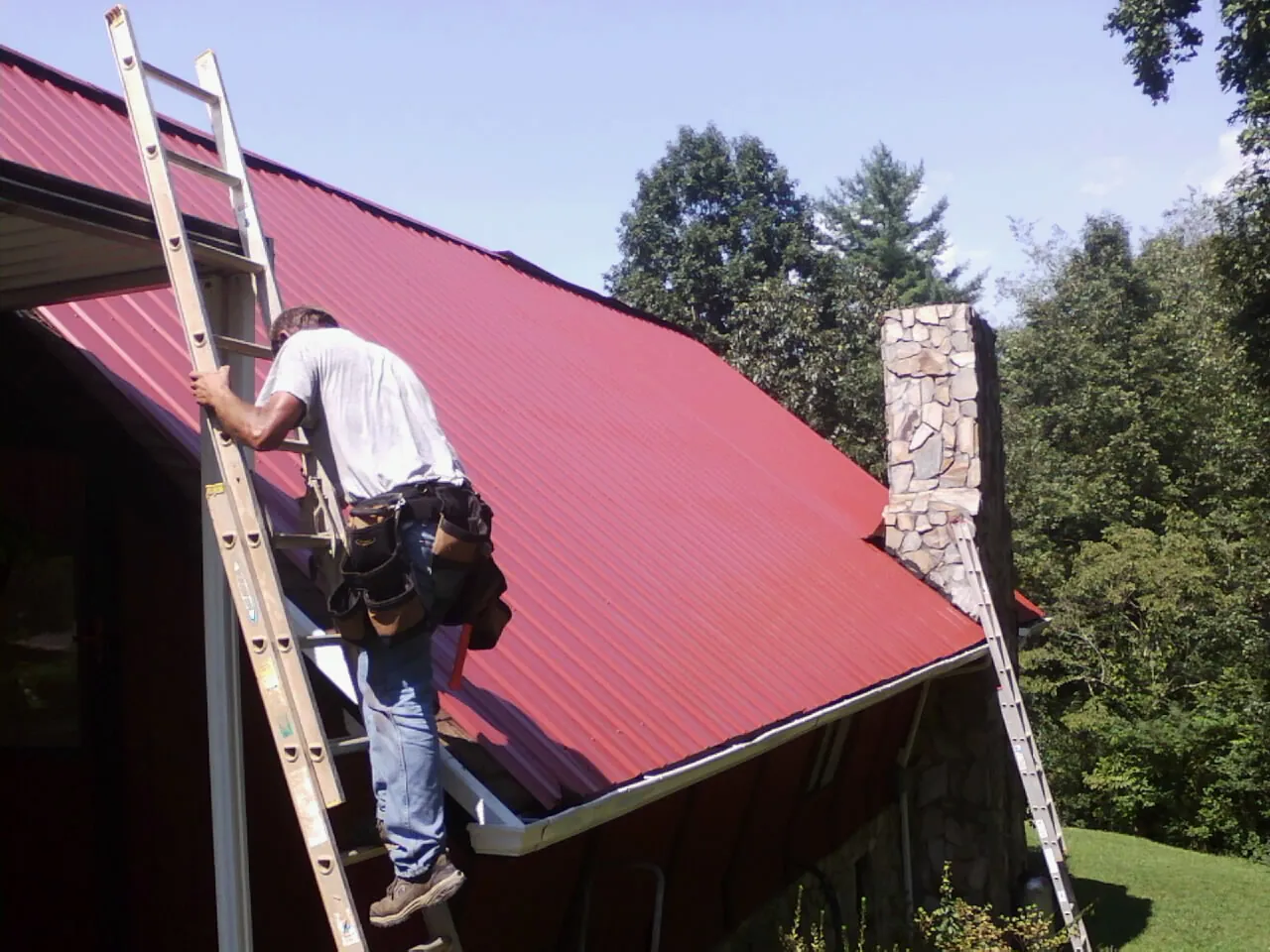 Roofer on a ladder beside a red metal roof and stone chimney — Soffit & Fascia Repair by Roofing Brothers in Mequon